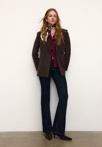 Rjavi kordurov dvoredni blazer, bordo bluza, temno širše kavbojke, rjavi koničasti škornji in vzorčast svileni šal. Enostavno ozadje.