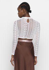 Weißes Spitzentop mit langen Ärmel, floralem Muster und Schlitzdetail am Rücken, kombiniert mit einem braunen Satinenrock mit Taillenband aus Satin.