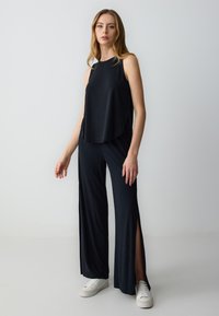 Top nero senza maniche con scollatura rotonda, abbinato a pantaloni larghi neri con fessure laterali. Indossato con sneakers bianche.