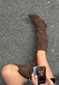 Botas de ante marrón hasta la rodilla con detalles de flecos, punta afilada y acabado texturizado, acompañadas de una mano sosteniendo un teléfono.