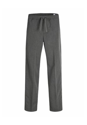 Garçon blond debout regardant vers la droite, portant un polo noir, un pantalon gris et des baskets blanches avec des bandes noires sur un fond uni.
