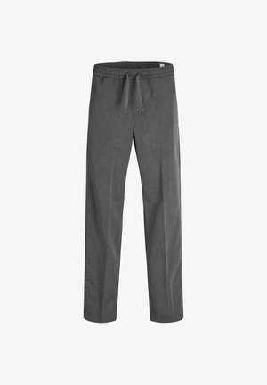 Garçon blond debout regardant vers la droite, portant un polo noir, un pantalon gris et des baskets blanches avec des bandes noires sur un fond uni.