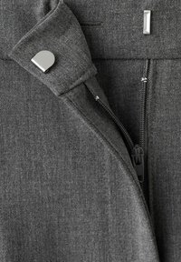 Tela gris oscuro con una cremallera parcialmente abierta y un botón a presión plateado en una solapa doblada, mostrando detalles de textura y costuras.