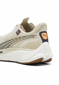 Ljusbeige löparskor med mesh-textur och vita detaljer. Har en gummisula och en marinblå logopatch på hälen.