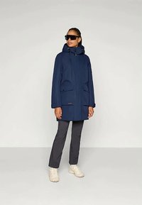 Nais is wearing a tume sinine kapuutsiga parka, tumedates hallides pükstes, valgetes sneakersides ja suurte mustade prillidega, seistes tasase heleda tausta ees.