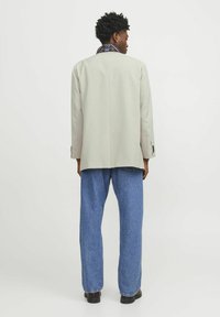 Blazer ajustado gris claro, llevado sobre una camisa de cuello negra y azul con patrón, combinado con jeans azul sueltos y zapatos oscuros.