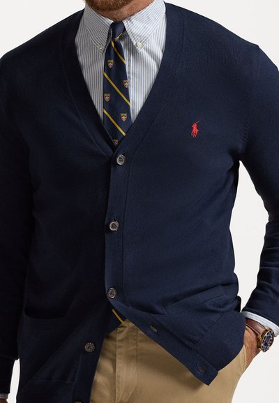 Cardigan bleumarin cu design în formă de v, închidere cu butoane și buzunare. Poartă peste o cămașă cu dungi bleu deschis și cravată cu model, având un logo roșu.