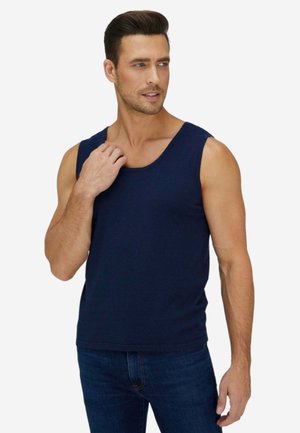 Mann trägt marineblaues ärmelloses Tanktop und dunkle Jeans, schaut leicht zur Seite, eine Hand nahe am Kragen.