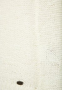 Pull blanc en tricot avec un motif texturé, orné d'un petit logo métallique près du bord inférieur.