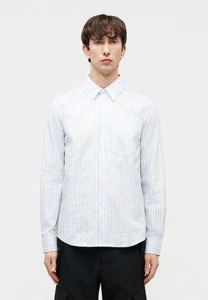 Νέος άνδρας που στέκεται κοιτώντας μπροστά, φορώντας ένα άσπρο πουκάμισο με γαλάζιες ρίγες και μαύρο παντελόνι, μπροστά σε απλό λευκό φόντο.