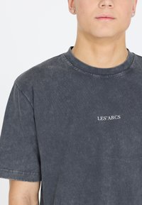 Nuori henkilö, joka on pukeutunut tummanharmaaseen kuluneeseen T-paitaan, jossa on valkoiset "LES' ARCS" -tekstit, ja pienet nappikorvakorut, kuvattu kaulasta keskivartaloon saakka.