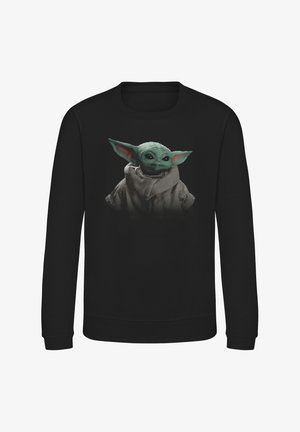 Sweatshirt noir avec un graphique d'un petit personnage à la peau verte, avec de grands yeux et des oreilles proéminentes, portant un manteau de couleur claire.