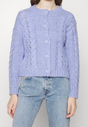 Vrouw draagt een lila grofgebreide cardigan met kabelpatronen en knopen, gecombineerd met lichtblauwe spijkerbroek.