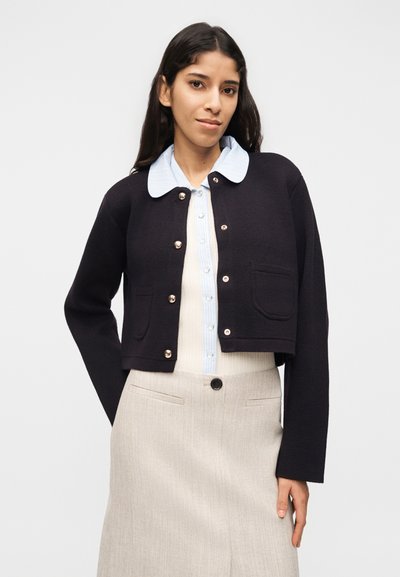 Γυναίκα που φοράει μαύρο κοντό ζακέτα με κουμπιά πάνω από μπλε ριγέ πουκάμισο με γιακά και μπεζ ψηλόμεση φούστα, σε απλό φόντο.