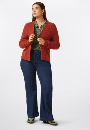 Rust-orange knitted cardigan with a textured diamond pattern, paired with a green patterned blouse and dark blue flared jeans.