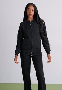 Casaco preto com fecho éclair e capuz, acompanhado de calças a condizer; feito de tecido suave; apresenta bolsos frontais e punhos canelados. Textura suave, design ajustado.