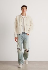 Chaqueta de denim beige claro sobre una camiseta blanca, combinada con jeans rotos de azul desgastado y zapatillas blancas. Diseño sencillo y ajuste relajado.