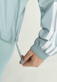 Lysblå atletisk jakke med elastiske manchetter, justerbart snor i kanten, med hvide striber langs ærmerne.