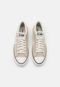 Beiga Converse All Star-sneakers med vita snören, vita gummitåspetsar och svarta detaljer, sedda ovanifrån på en vit bakgrund.