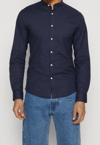 Maglietta a maniche lunghe blu navy con colletto alla cinese, chiusura a bottoni e una texture liscia, abbinata a pantaloni di denim azzurro chiaro.