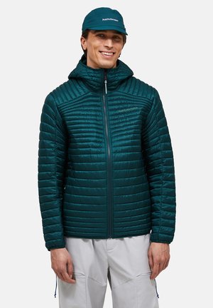 Man draagt een donker petrolkleurige doorgestikte jas met capuchon en bijpassende merkpet, staat en glimlacht tegen een effen achtergrond.