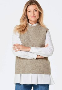 Beige gestrickte Sweaterweste mit hohem Kragen, getragen über einem weißen Hemd mit Kragen. Die Weste hat eine gerippte Textur und eine lockere Passform.