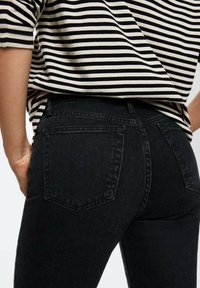 Jean en denim noir à coupe ajustée, avec deux poches arrière et une taille haute. Porté avec un haut à manches courtes rayé.