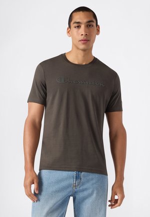 Junger Mann trägt ein dunkelbraunes Champion T-Shirt und hellblaue Jeans und steht vor einem einfarbigen weißen Hintergrund.
