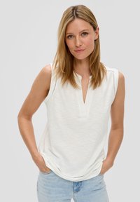 White sleeveless top with a notch collar, textured fabric, decorative stitching at shoulders, and a relaxed silhouette. Paired with light blue jeans.