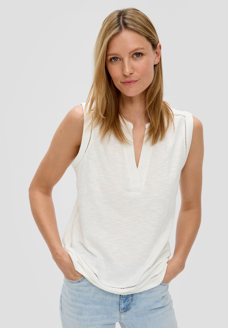 White sleeveless top with a notch collar, textured fabric, decorative stitching at shoulders, and a relaxed silhouette. Paired with light blue jeans.