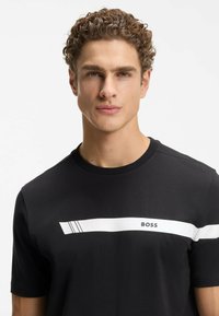 Schwarzes Baumwoll-T-Shirt mit weißem horizontalem Streifen, der aus drei Linien besteht, und dem Schriftzug "BOSS" auf der Brust. Kurze Ärmel, Rundhalsausschnitt.