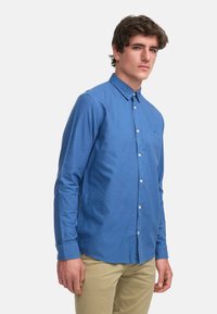 Camisa azul de botones, hecha de algodón, con mangas largas y cuello. Presenta un diseño liso y un sutil logo bordado en el pecho.
