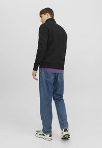 Felpa nera con zip e collo alto, abbinata a una maglietta viola a strati e jeans blu a vestibilità ampia. Sneakers bianche con dettagli neri.