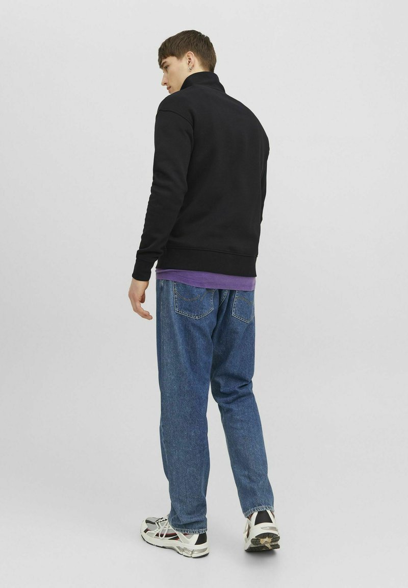 Zwarte zip-up sweatshirt met hoge kraag, vergezeld van een gelaagd paarse shirt en blauwe, loszittende jeans. Witte sneakers met zwarte accenten.