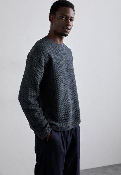 Sweatshirt texturé gris foncé avec un motif tissé, encolure ronde et poignets côtelés, associé à un pantalon ample et sombre.