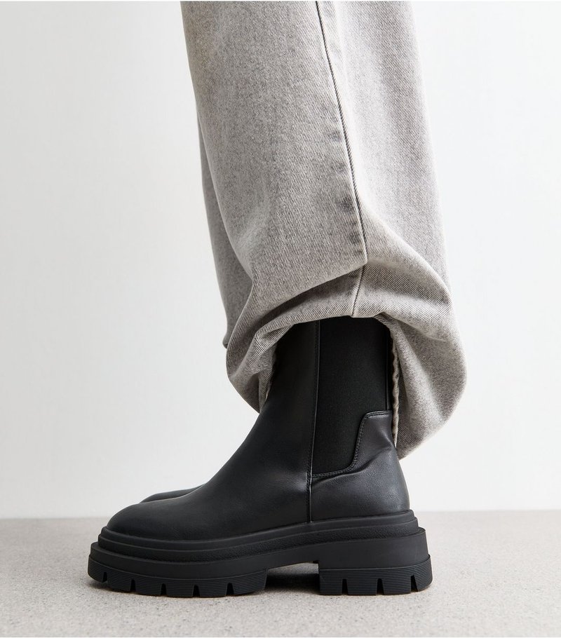 Bottes en cuir à semelle épaisse noires portées avec un pantalon en denim gris clair lâche sur un sol clair et un fond uni.