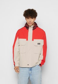 Jachetă color-block în roșu și bej, cu glugă, două buzunare mari în față, închidere cu botnițe și manșete elastice. Fabricată dintr-un material ușor.