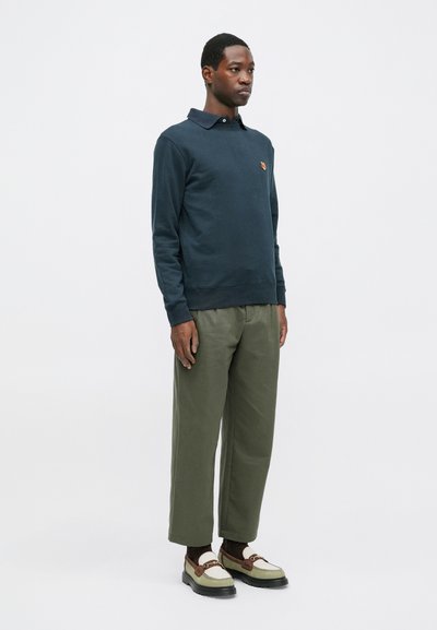 Hanorac bleumarin cu guler și un mic logo portocaliu, asortat cu pantaloni largi de culoare verde măsliniu și încălțăminte bicoloră.
