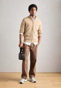 Beige glidelås strikkegenser, hvit skjorte med krage, brune bukser, svart teksturert veske, hvite og grå sneakers, stående på tregulv.