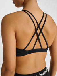 Frau trägt einen schwarzen Sport-BH mit dünnen, gekreuzten Rückenriemen und schwarze Sportleggings mit weißem Markenlogo am Bund.