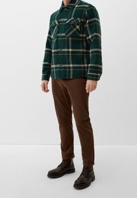 Giacca a scacchi verdi con due tasche sul petto, bottoni marroni e colletto, abbinata a pantaloni marroni e stivali neri con lacci.