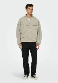 Giacca a zip in pile di un beige chiaro con cuciture scure, colletto alto e tasche laterali, abbinata a pantaloni neri e sneakers nere.