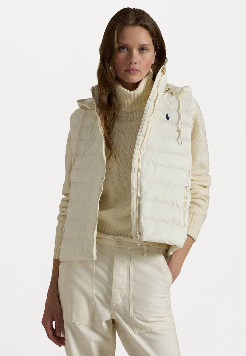 Puffige cremefarbige Weste mit Reißverschluss vorne, Kapuze und seitlichen Taschen. Getragen über einem strukturierten cremefarbenen Rollkragenpullover und passenden cremefarbenen Hosen. Feine Logodetails.