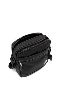 Čierna crossbody taška vyrobená z hladkej látky, obsahujúca dva zipsové oddiely, nastaviteľný popruh a malú nášivku s logom na prednej strane.
