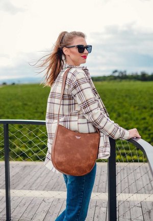 Brown faux leather shoulder bag with a curved shape and logo, worn over a plaid shirt, against a green field backdrop.
