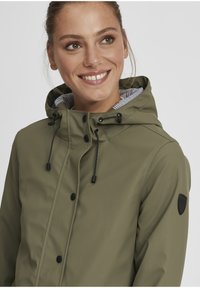 Veste imperméable vert olive avec capuche, dotée de boutons noirs, de cordons de serrage et d'une doublure intérieure rayée. Comprend un patch logo sur la manche.