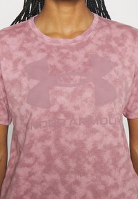 Růžové tričko s krátkým rukávem s texturovaným, vybledlým vzorem. Má velké, tónované logo Under Armour a text na spodní části.