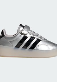 Srebrna tenisica s crnim prugama od patentne kože, teksturiranim srebrnim gornjim dijelom i prozirnim potplatom. Ima kopču na čičak i vezice.