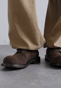 Zapatos de fieltro marrón con una puntera redondeada y una suela oscura y lisa. Cuentan con cordones y un sutil detalle de costura decorativa.