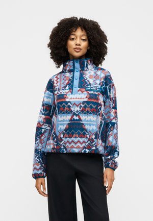 Mujer con cabello rizado que lleva un jersey de forro polar con estampado en azul, rojo y blanco con botones a presión y pantalones negros, de pie frente a un fondo blanco.